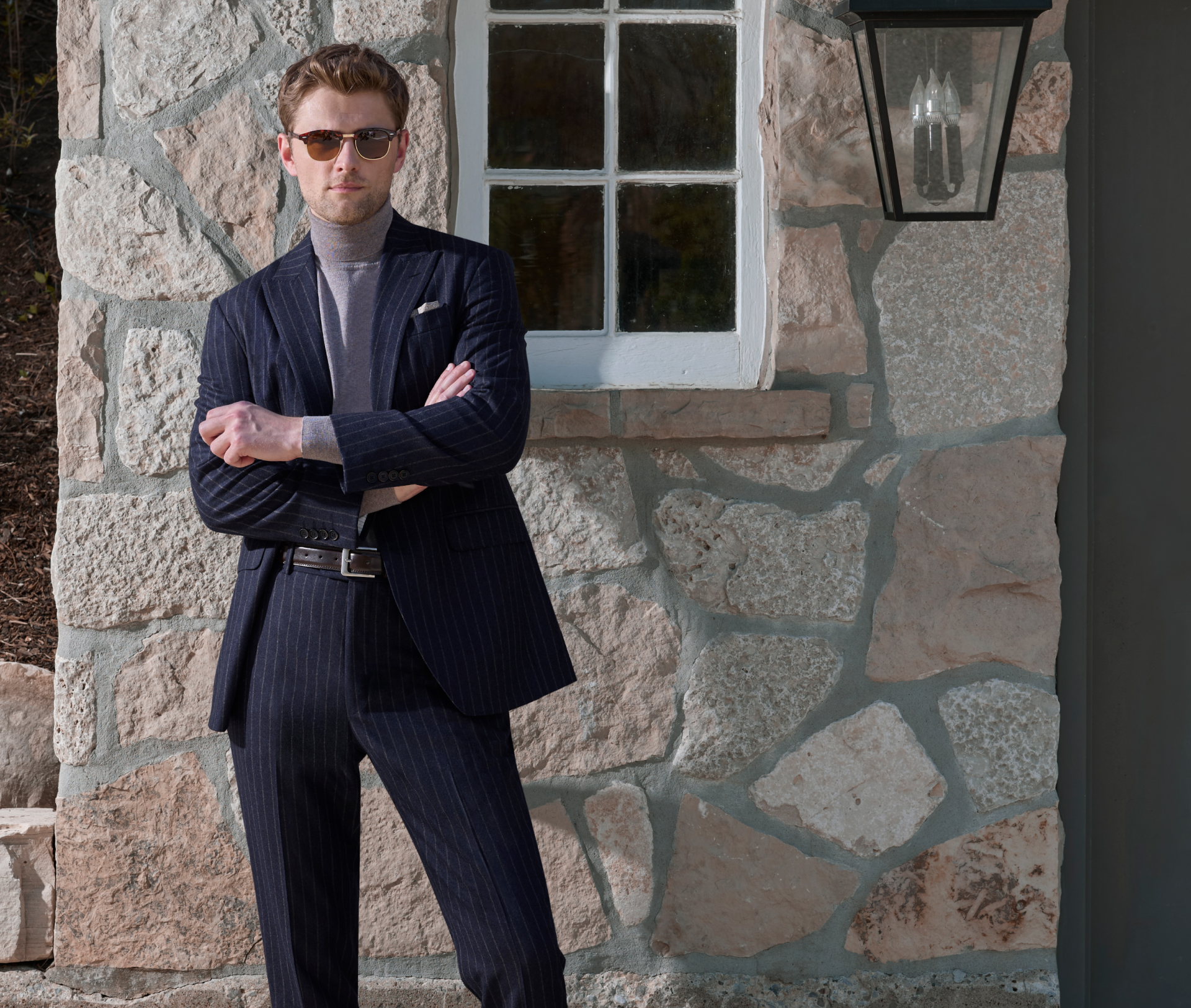 A young man wearing a navy flannel suit with a subtle brown stripe stands confidently in front of a white window beside a vintage lamp and a grey door set into a stone wall. He has his arms crossed and is wearing sunglasses.