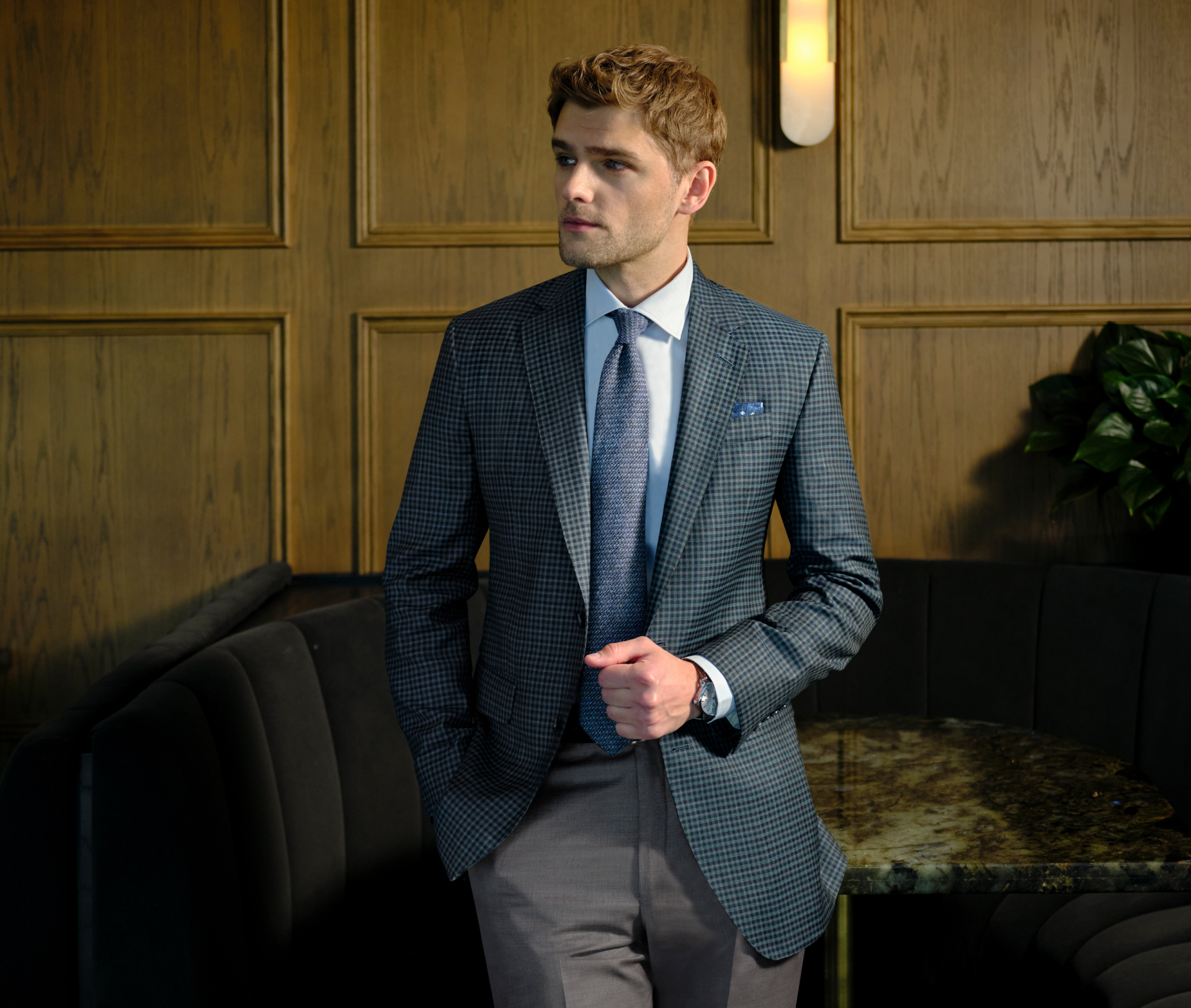 A young man leans against a table wearing a green, navy, and grey micro-check sport coat with tailored trousers. He looks into the distance while holding the front of his jacket with one hand and keeping the other in his trouser pocket. One leg is crossed over the other. Behind him is a vintage-style wall with a subtle plant in the corner.