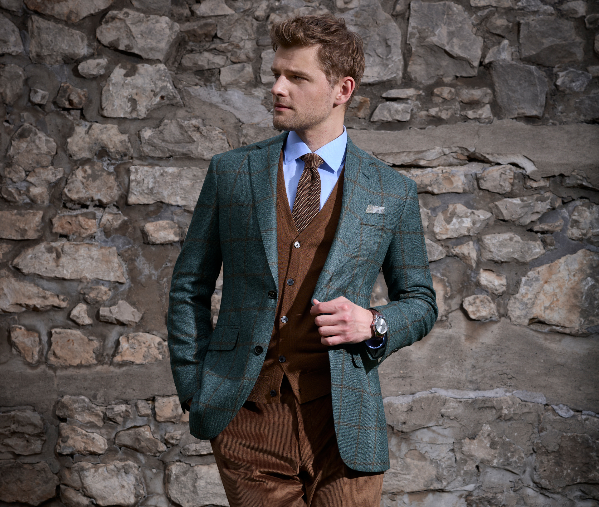 A young man wearing a retro styled teal sport coat and brown trousers stands in the grass with his leg crossed, with one hand in his pocket and the other adjusting his jacket while looking to the side.