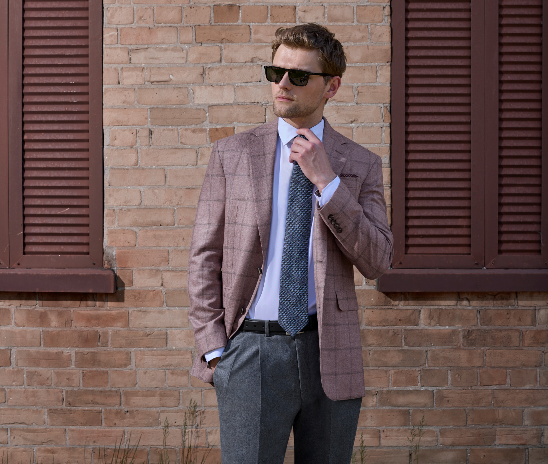 A young man wearing a soft pink jacket with a grey windowpane pattern stands on the grass in front of a light yellow brick wall with burgundy shutters. He adjusts his tie with one hand while the other rests in his pocket as he wears black sunglasses.