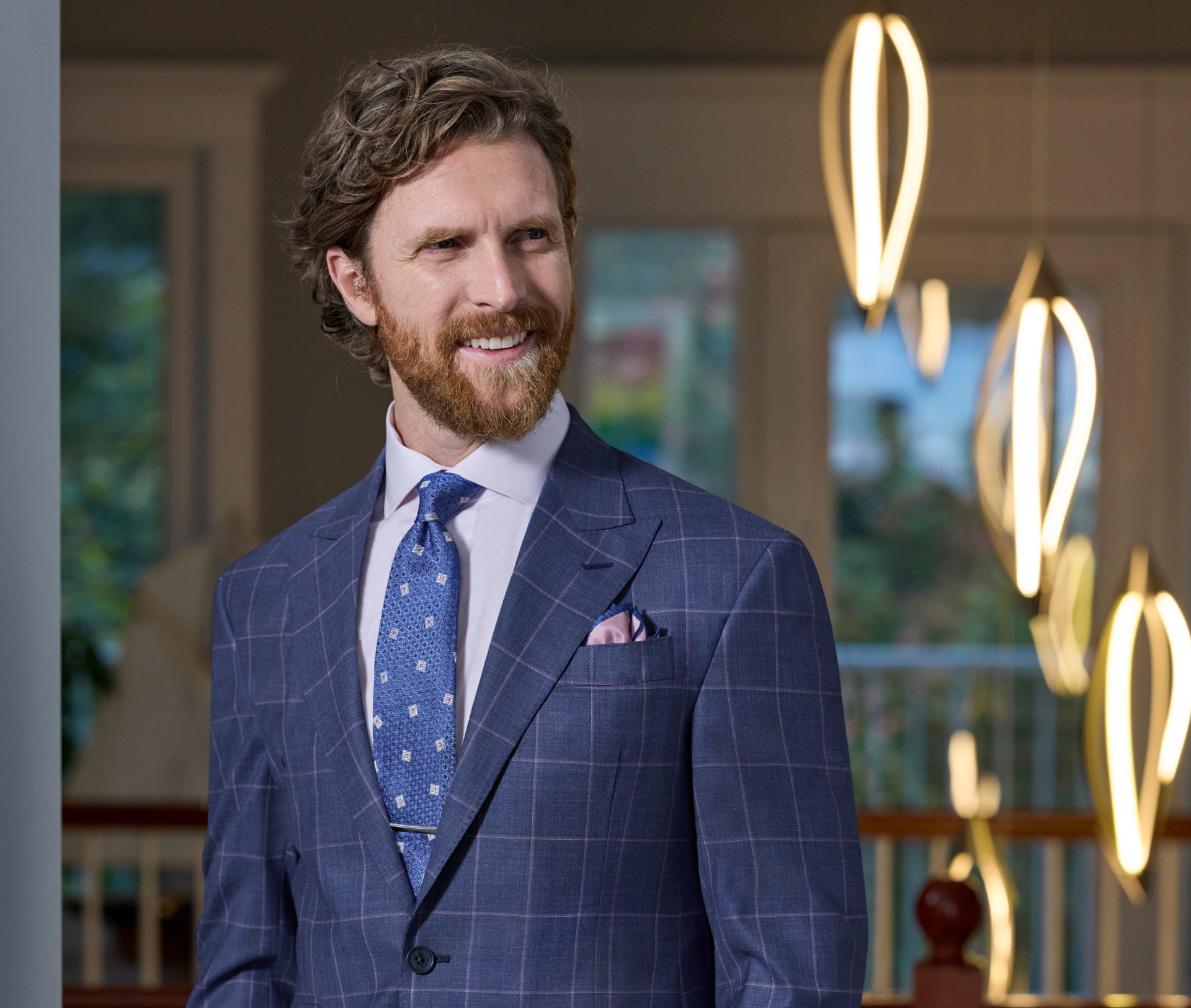 A smiling bearded man is wearing a blue and purple windowpane suit with a peak lapel. He is standing in front of a banister and ceiling lights with his hands in his pockets.