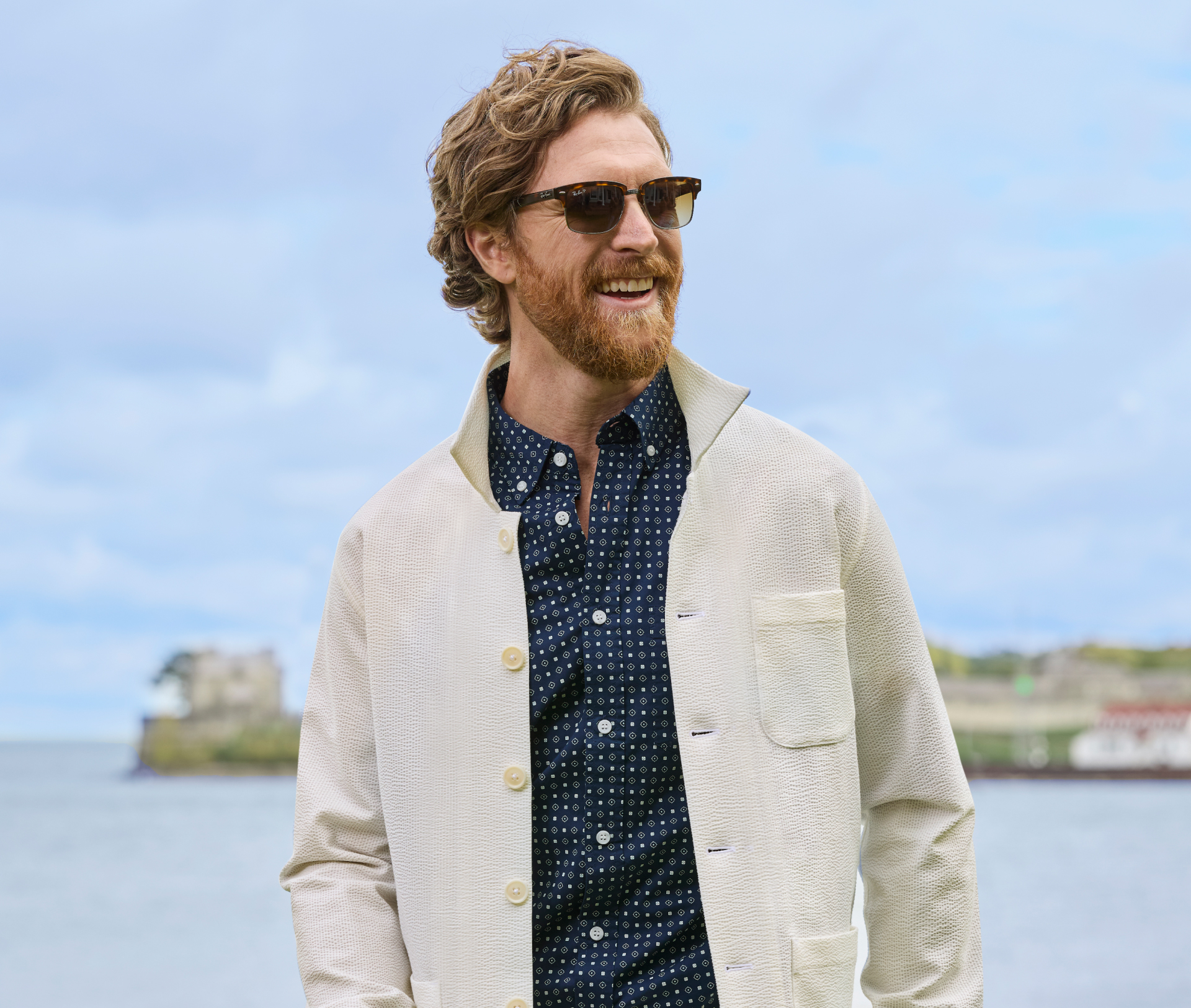 A smiling bearded man is wearing a cream shirt jacket and trouser made of a cotton stretch seersucker. He is wearing sunglasses and is walking outside with a lake behind him.