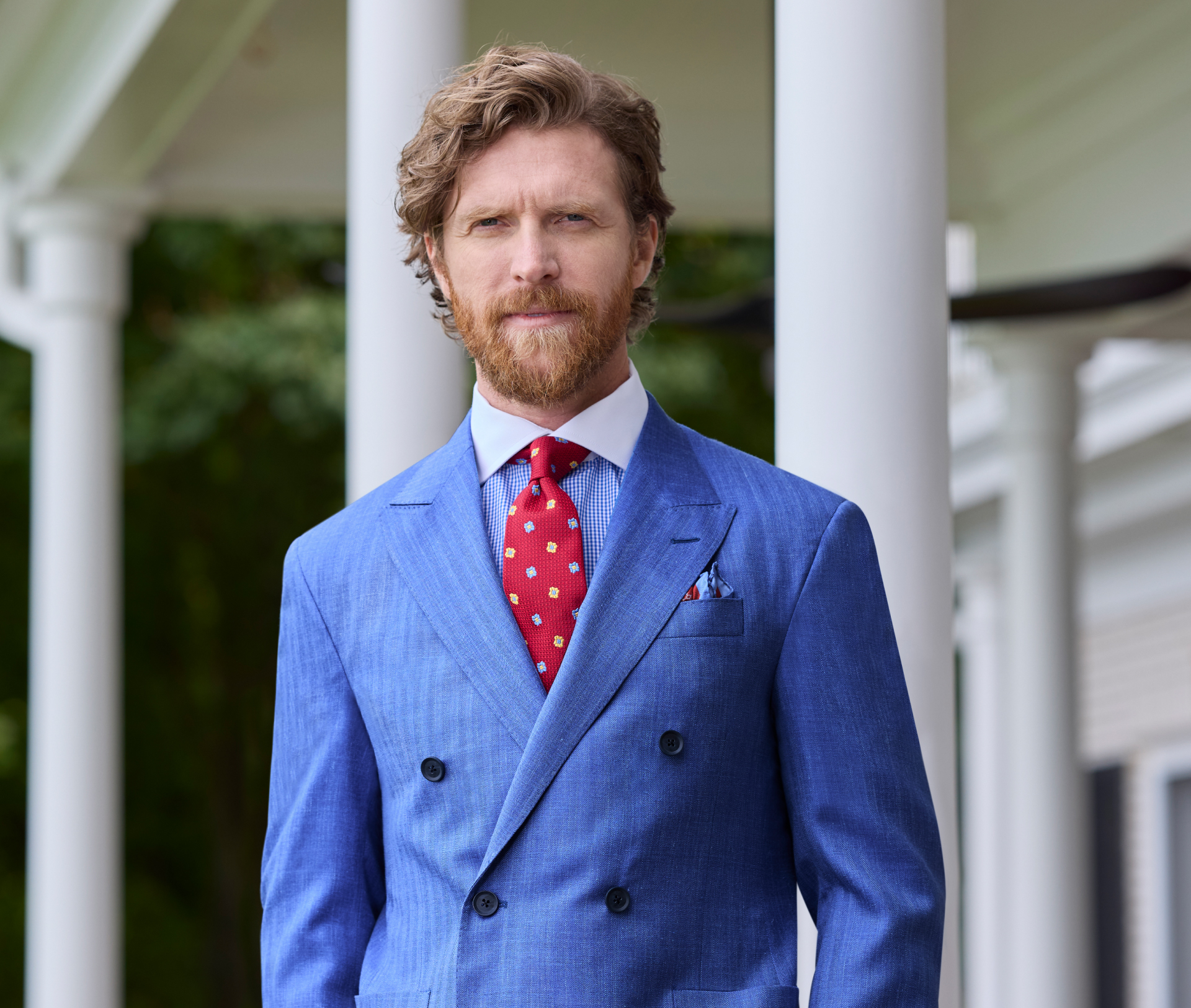 A bearded man is wearing a blue herringbone double-breasted sport coat paired with cream trousers. He has his hands in his pockets and is standing in front of white columns. He is also wearing a bold red patterned necktie.