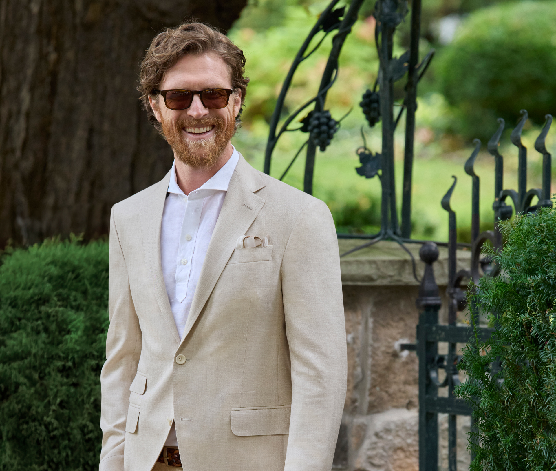 A smiling bearded man is wearing a beige herringbone two-button single-breasted suit with a notch lapel. He is standing outside in front of a gate with bushes nearby. He is also wearing sunglasses and has his right hand in his pocket.